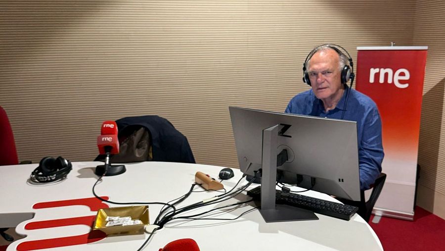 Periodista en estudio de radio con ordenador, auriculares y micrófonos.  Mesa blanca, suelo rojo y logo RNE visible.