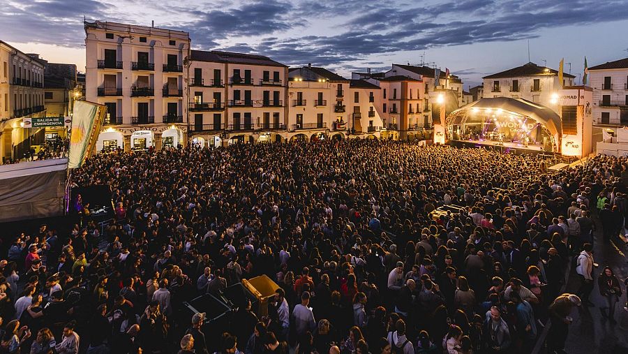 Concierto nocturno en una plaza con multitud de asistentes, escenario iluminado y equipos de sonido visibles.