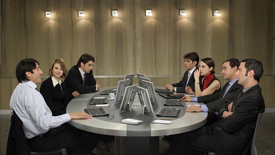 Siete personas alrededor de una mesa ovalada con ordenadores, en una sala con paneles de madera.  Ambiente serio y profesional.