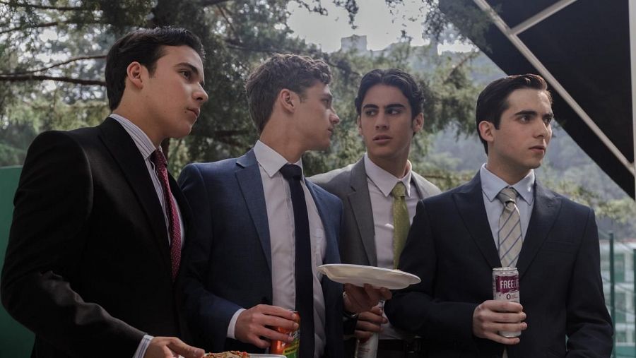 Cuatro hombres en trajes y corbatas conversan informalmente al aire libre, sosteniendo comida y bebida.  Se observa vegetación y una construcción en el fondo.