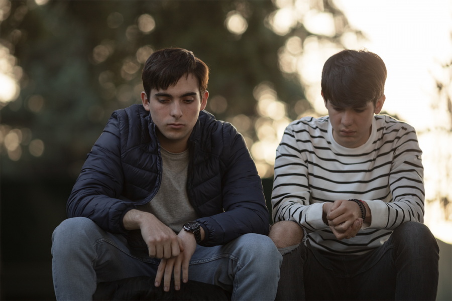 Dos chicos jóvenes, uno con chaqueta azul y otro con jersey a rayas, sentados en un parque con expresión seria. Sus posturas sugieren introspección.