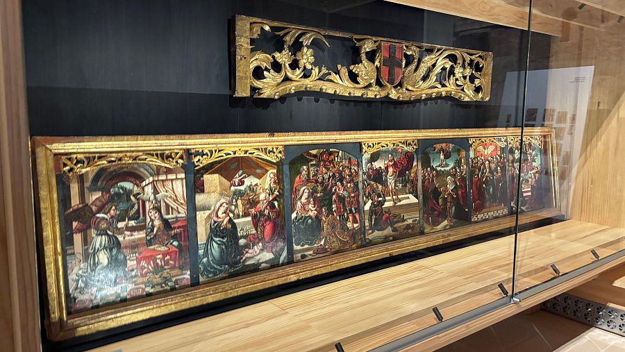 Predela de un retablo religioso en vitrina, con paneles que muestran escenas religiosas y decoración dorada.  Sobre ella, otra pieza con decoración dorada y un escudo.