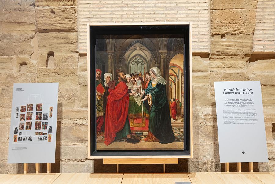 Pintura renacentista de la Presentación de Jesús en el Templo, expuesta en un atril. La Virgen entrega al niño a un hombre con túnica roja; otros personajes observan.  Carteles informativos acompañan la obra.
