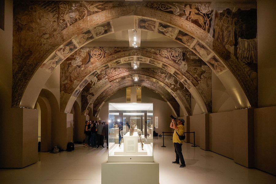 Pintura mural en tonos terrosos en techos abovedados de una sala de museo. Vitrina central con objetos expuestos y visitantes observando.