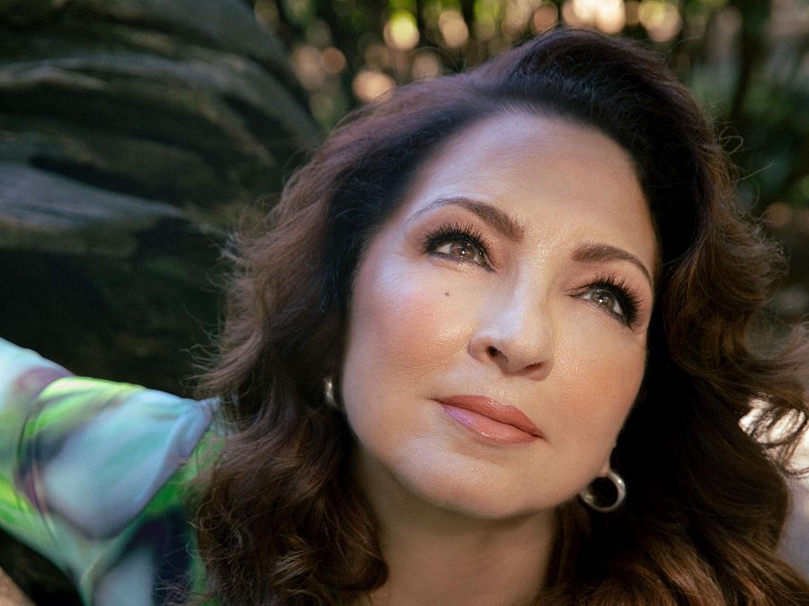 Gloria Estefan, con blusa estampada y pendientes, mira hacia arriba con expresión serena.  Imagen que acompaña una noticia sobre la celebración de las raíces latinas.