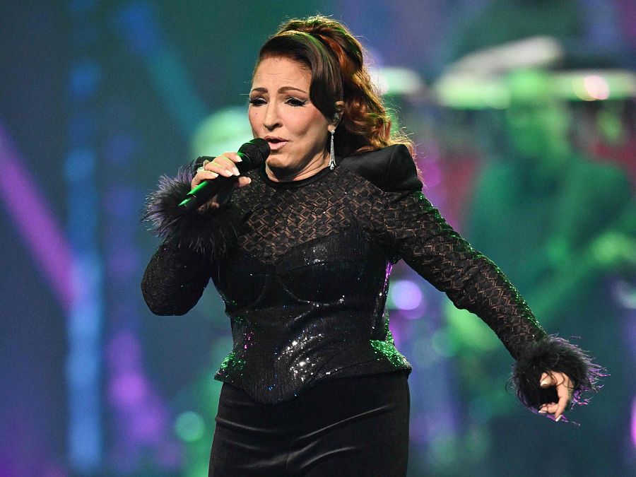 Cantante con traje negro brillante y plumas actuando en un escenario iluminado.  Sujeta un micrófono y mira al público.