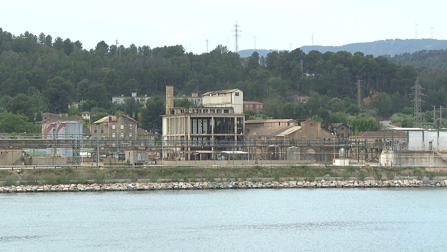 Vista del río Ebro, tranquilo, con una zona industrial y una planta química al fondo.  Se aprecian rocas en la orilla y edificios de diversos tamaños y estados de conservación.