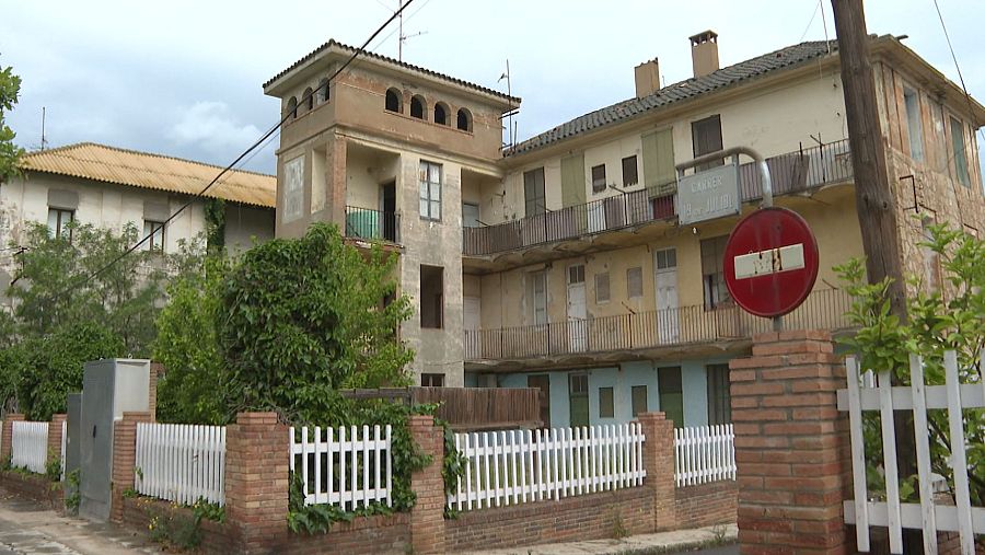 Dos edificios deteriorados de una colonia industrial, uno con torre y arcos, el otro con tejado de tejas, separados por una valla con señal de prohibido el paso.