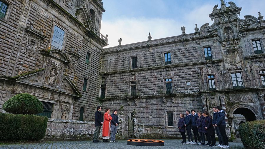 Grabación de un programa de televisión frente a un monasterio. Dos presentadores y un grupo de aspirantes posan para la cámara, con una marca circular en el suelo.