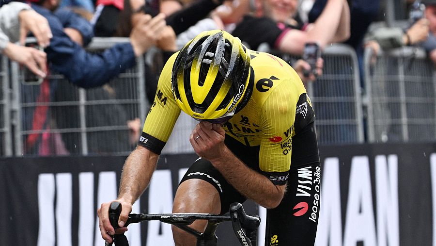 Ciclista con maillot amarillo, cabeza agachada y mano en la cara, mostrando gran emoción tras cruzar la meta. Público visible al fondo.