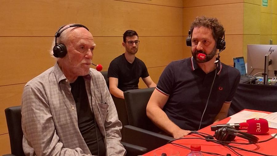 Grabación en estudio con tres hombres; dos con auriculares y micrófonos (uno con logo RNE) sentados a una mesa con un portátil HP; el tercero observa.