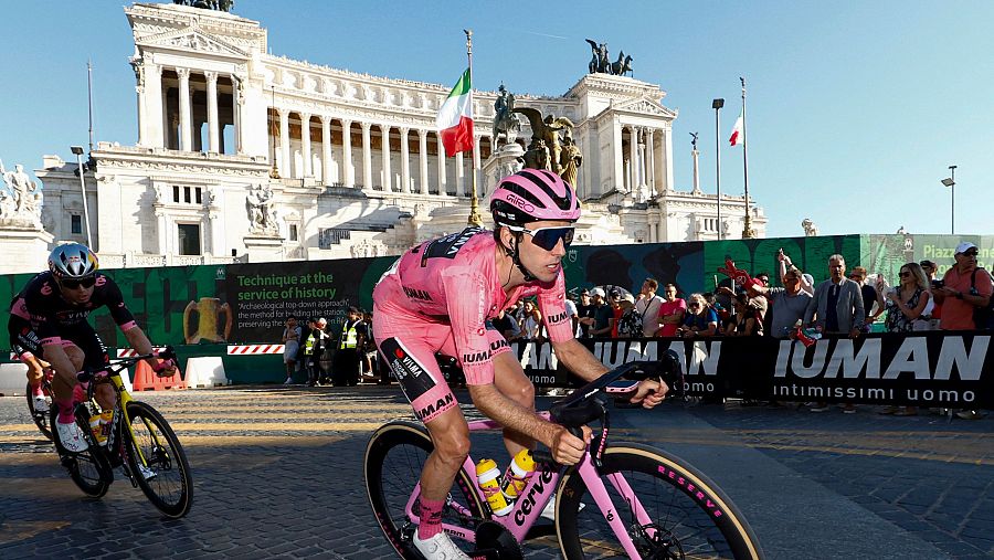 Ciclista con maglia rosa en carrera, posiblemente cerca de la meta, con el Monumento a Víctor Manuel II al fondo y público presente.