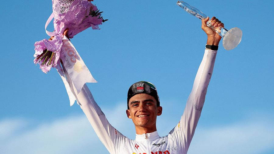 Ciclista con maillot blanco celebra victoria, sosteniendo flores y trofeo. Sonrisa de alegría, cielo azul de fondo.