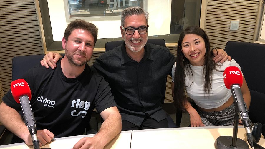 Foto de tres personas sonriendo en un estudio de radio. El presentador central lleva camisa oscura y gafas; los acompañantes, camisetas informales. Dos micrófonos con logo son visibles.
