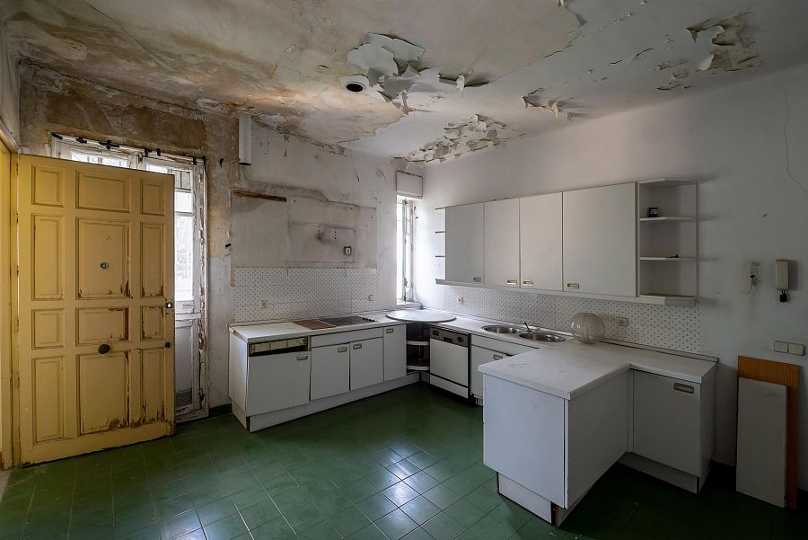 Cocina abandonada con muebles blancos antiguos, puerta amarilla deteriorada y suelo de baldosas verdes.  Evidentes signos de deterioro y humedad.