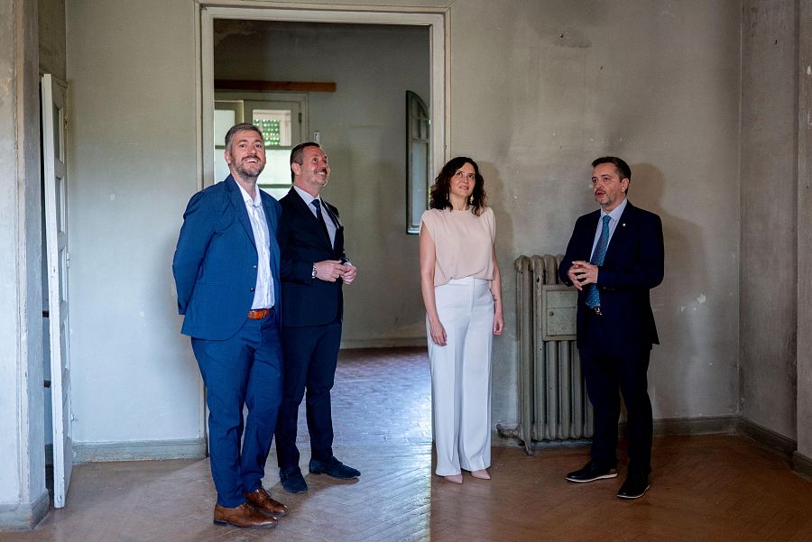 En una habitación con paredes sin pintar y suelo de madera, cuatro personas, con vestimenta formal, observan algo fuera de la imagen.