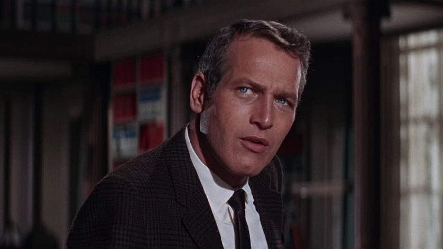 Paul Newman como Harper, investigador privado, con cabello rubio grisáceo peinado hacia atrás, viste chaqueta oscura a cuadros sobre camisa blanca y corbata, expresión seria.