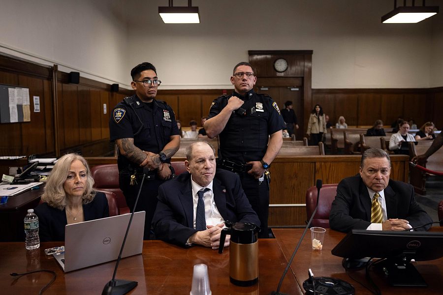 En una sala de tribunal, Harvey Weinstein, en traje, se sienta en una mesa con micrófonos, junto a una mujer y un hombre en traje, y dos policías de pie detrás.