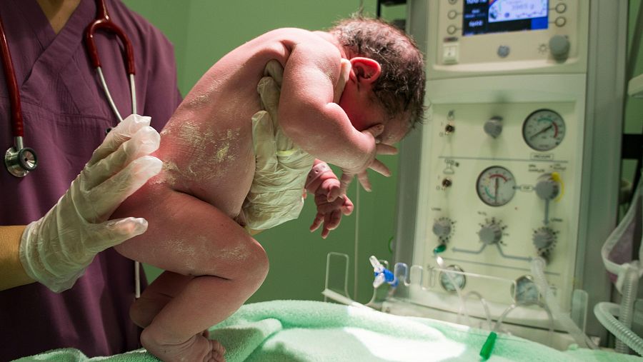 Recién nacido cubierto de vérnix, sostenido por un profesional sanitario con uniforme morado y guantes, cerca de equipo médico para monitorización.