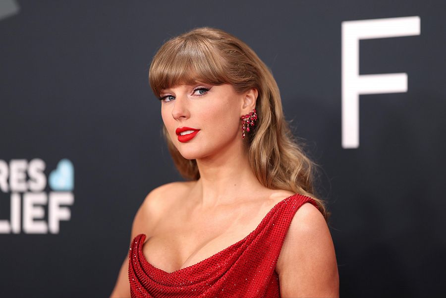 Taylor Swift con vestido rojo, un solo tirante y pendientes rojos en una ceremonia con fondo oscuro y una gran 'F' blanca visible.
