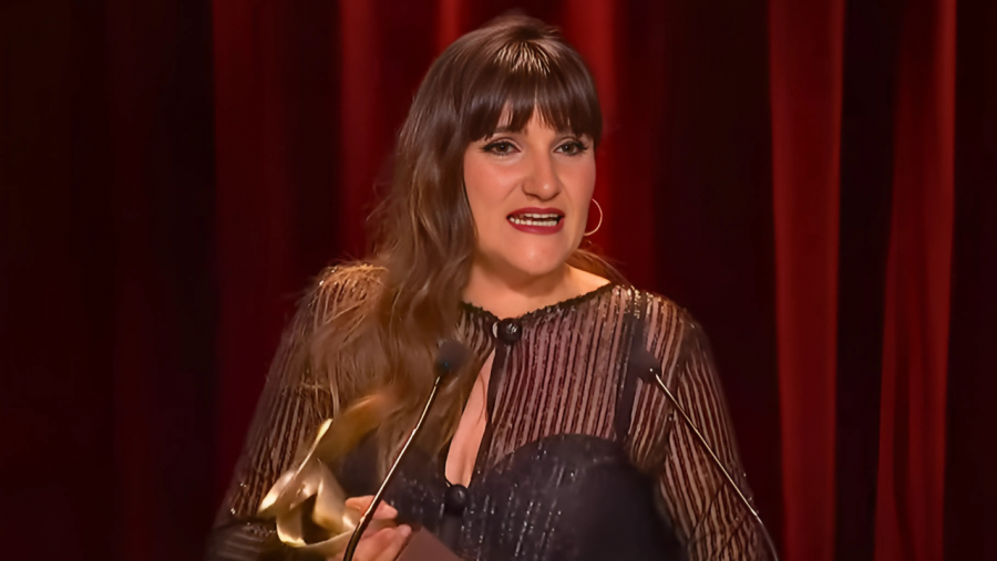 Cantante con vestido oscuro y pendientes recibe premio dorado en escenario con telón rojo.  Muestra una expresión de felicidad.