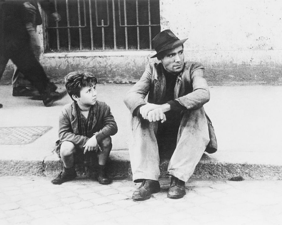 Dos personas, un hombre y un niño, sentados en un bordillo con expresiones serias. Fondo con peatones y una reja.