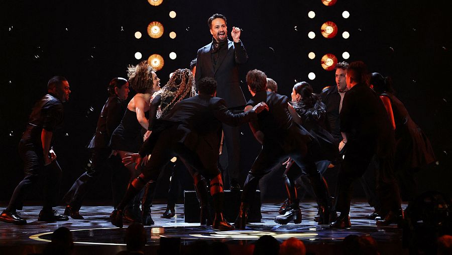 El elenco de Hamilton y Lin-Manuel Miranda, en escena con atuendos oscuros, durante una actuación en los Premios Tony. Iluminación cálida y dramática.