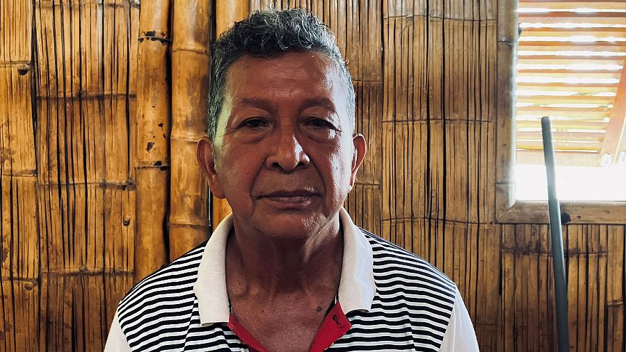 Hombre mayor con polo a rayas horizontales, sentado frente a pared de bambú.  Luz natural entra por ventana con persiana de bambú.