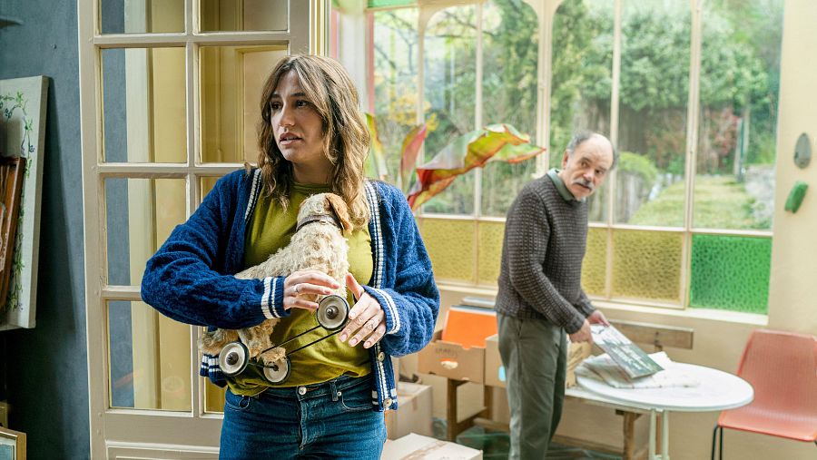 Mujer con juguete antiguo y expresión pensativa; al fondo, hombre con discos de vinilo.  Escena en interior con cajas, posible mudanza o reorganización.