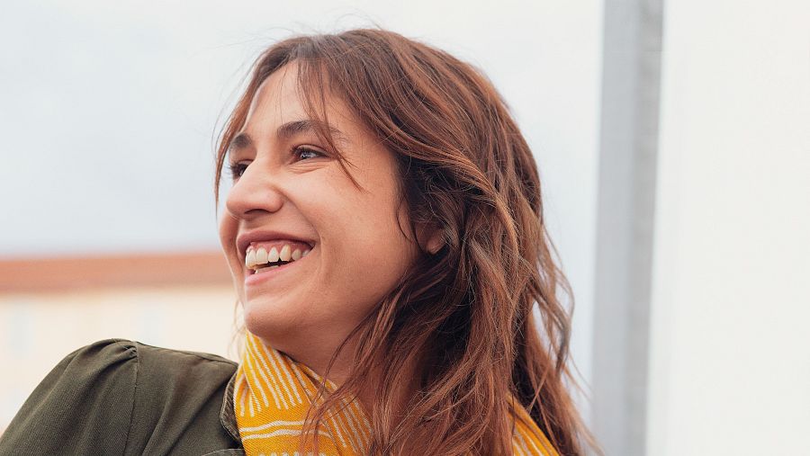 Joven de cabello castaño claro y ondulado, con chaqueta verde oliva y pañuelo amarillo a rayas, sonríe mirando hacia arriba. Fondo desenfocado con edificio anaranjado.