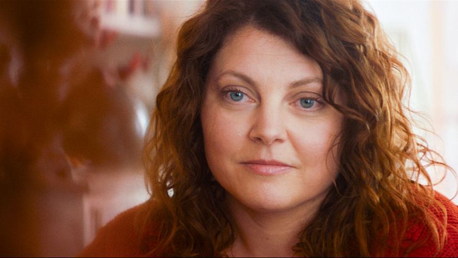 Mujer con cabello rojizo y rizado, ojos azules y expresión seria, mirando hacia la izquierda. Lleva una prenda roja. Fondo desenfocado, interior.