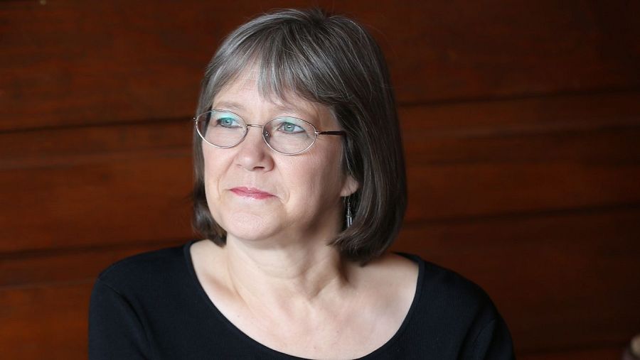 Retrato de una mujer de edad madura con pelo corto canoso y gafas, vistiendo camisa oscura, con expresión pensativa ante un fondo de madera.