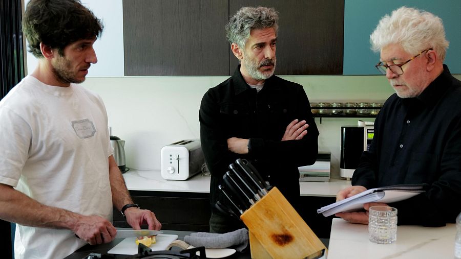 El director Pedro Almodóvar instruye a dos actores en una cocina; uno observa atento, el otro corta algo en una tabla.  Se ven electrodomésticos y un guion.