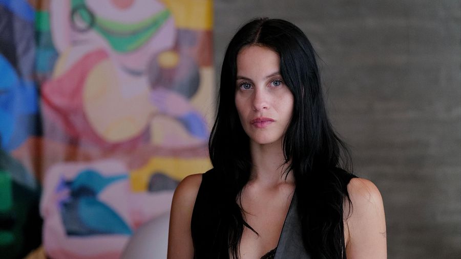 Milena Smith, con top negro, mira a cámara en una escena de rodaje.  Fondo con pintura abstracta vibrante.