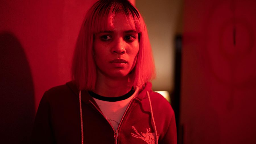 Mujer joven con pelo rosa y sudadera granate, mirada seria a cámara, en espacio rojo oscuro.