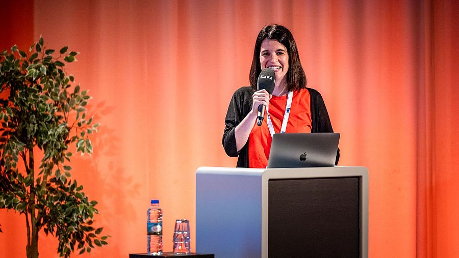Presentadora de Radio Clásica hablando al micrófono durante un evento de noticias culturales. Lleva camisa naranja y chaqueta negra; el atril tiene un portátil.