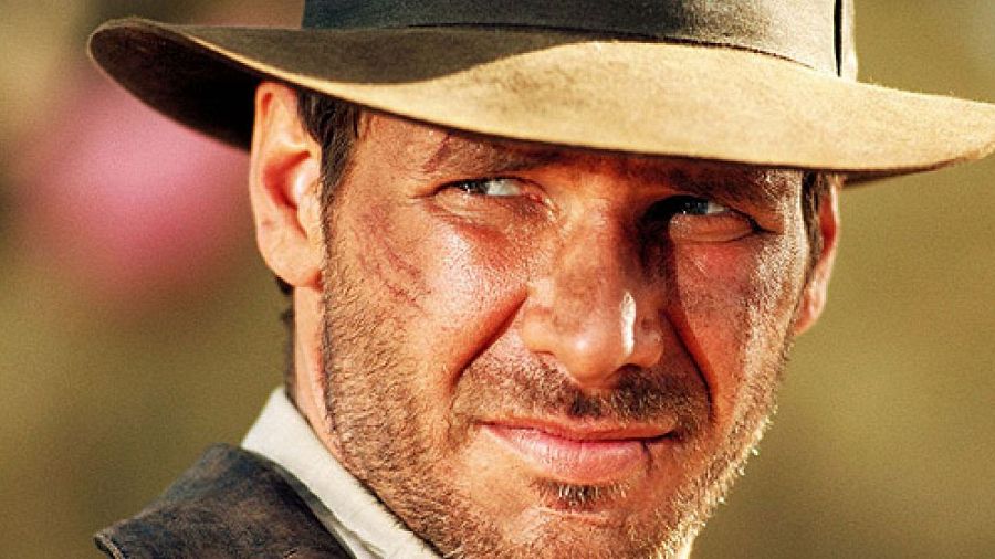 Primer plano de Harrison Ford con su característico sombrero, mostrando una leve sonrisa; evoca la imagen de Indiana Jones.