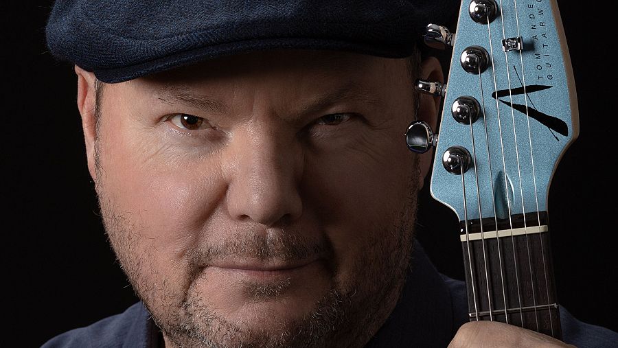Músico con gorra azul marino y barba, sosteniendo una guitarra eléctrica azul clara. 