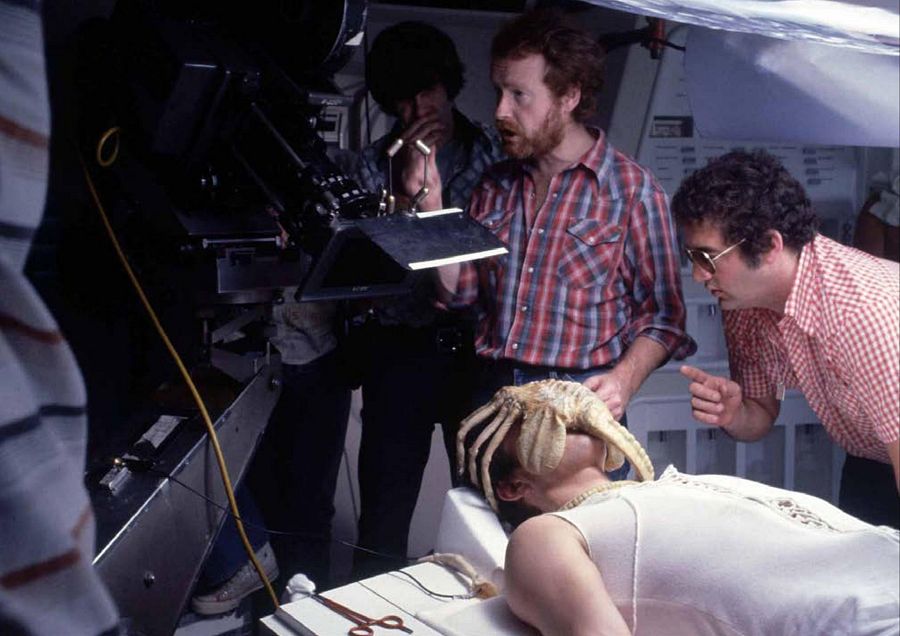 El director Ridley Scott, con camisa a cuadros, dirige una escena de rodaje con un actor y un *facehugger*. Se observan tijeras y una cámara en el set.
