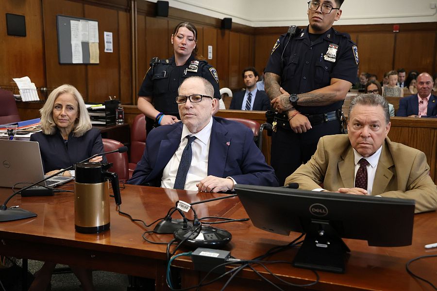 Sala de tribunal con Harvey Weinstein y otros individuos sentados a una mesa con micrófonos, un ordenador y una jarra. Agentes de policía presentes.