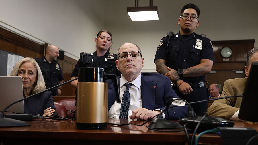 Imagen de Harvey Weinstein en una sala judicial, vestido formalmente, esperando un veredicto.  Presencia policial visible.