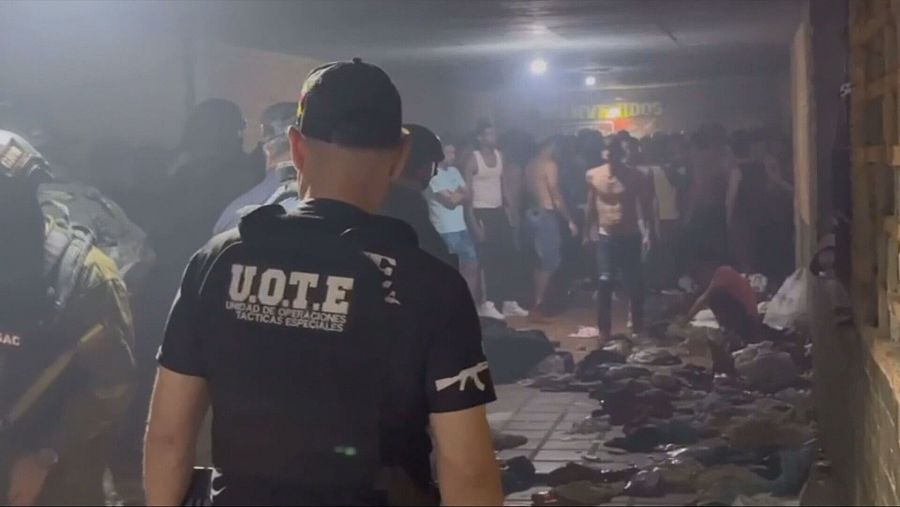Agente de la UOTE, con uniforme negro y gorra, en pasillo de cárcel venezolana.  Escena con poca luz, personas y objetos en el suelo, indicando posible operativo policial.