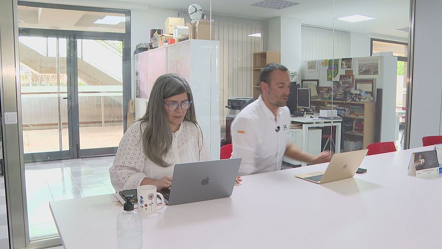 Dos personas en una oficina, una mujer trabajando en un portátil y un hombre junto a otro portátil.  Mesa con objetos y fondo con estanterías.