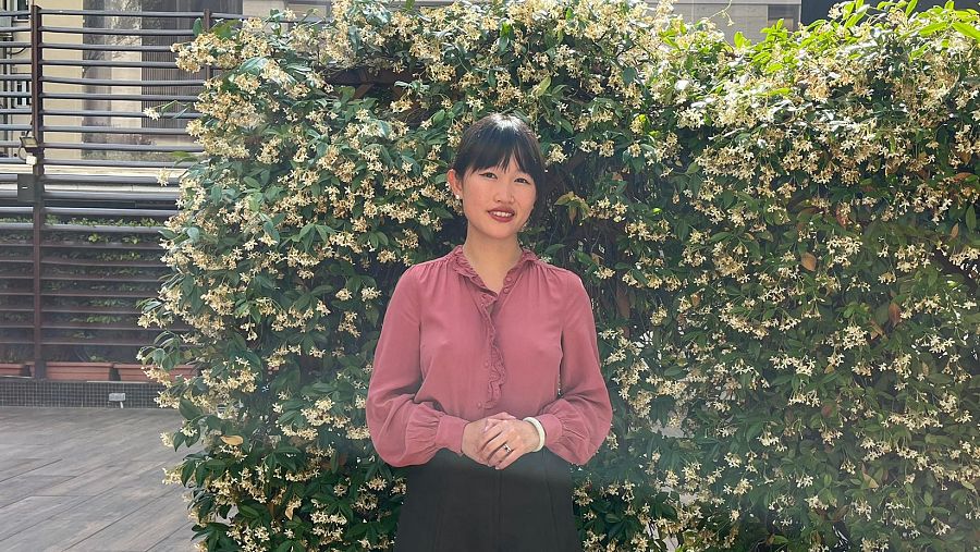 R. F. Kuang en entrevista: blusa rosa, pantalones negros, pulsera blanca. Fondo con planta floreciente y celosía.