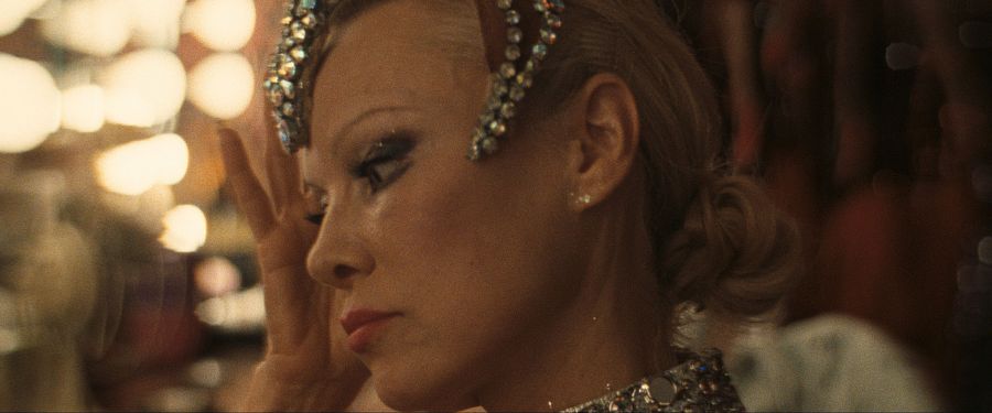 Mujer con tocado y maquillaje de escena, preparándose entre bastidores.  Expresión seria y concentrada. Fotograma de película.