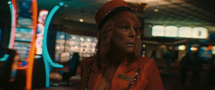 Mujer pelirroja mayor, con gorra y chaqueta rojas, en un ambiente con luces de neón rojas y turquesas.  Expresión seria.