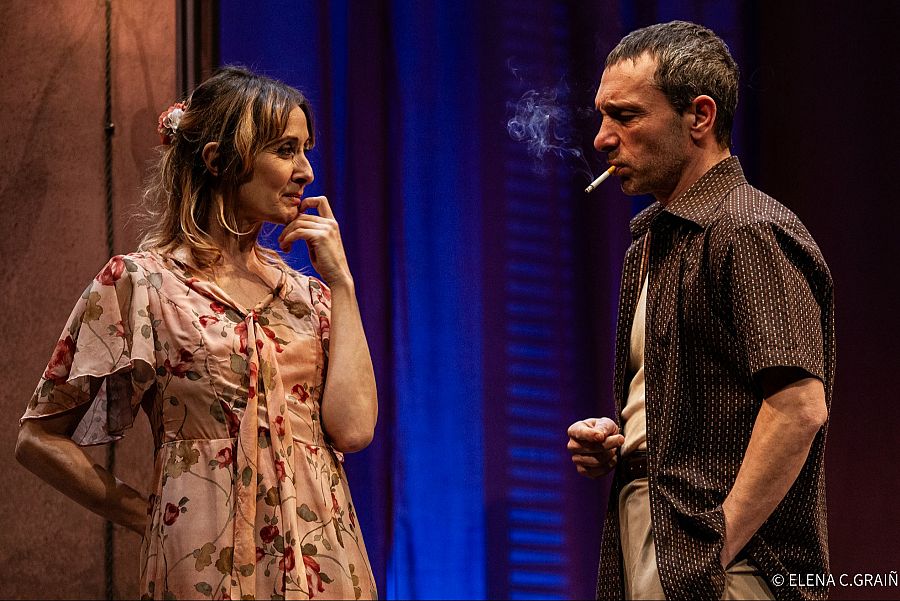 Escena teatral: Blanche (vestido rosa) con expresión pensativa y Stanley (camisa oscura, cigarrillo) con mirada seria.  Iluminación y fondo azul oscuro crean atmósfera de tensión.
