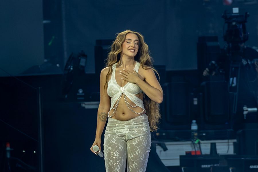 Cantante con atuendo blanco actuando en un escenario, micrófono en mano y expresión sonriente. Se observa equipo de sonido y cámara.