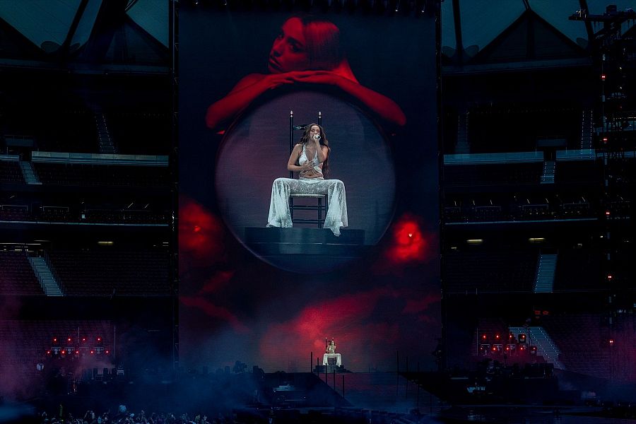Cantante en concierto, sentada en silla con micrófono, vestida de blanco sobre escenario con iluminación roja y humo. Gran pantalla con imagen ampliada de la artista.