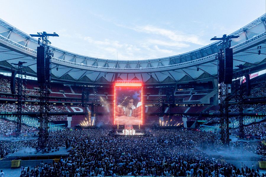 Concierto multitudinario en estadio; escenario con bailarines y pantalla gigante mostrando a la artista; iluminación blanca y roja.